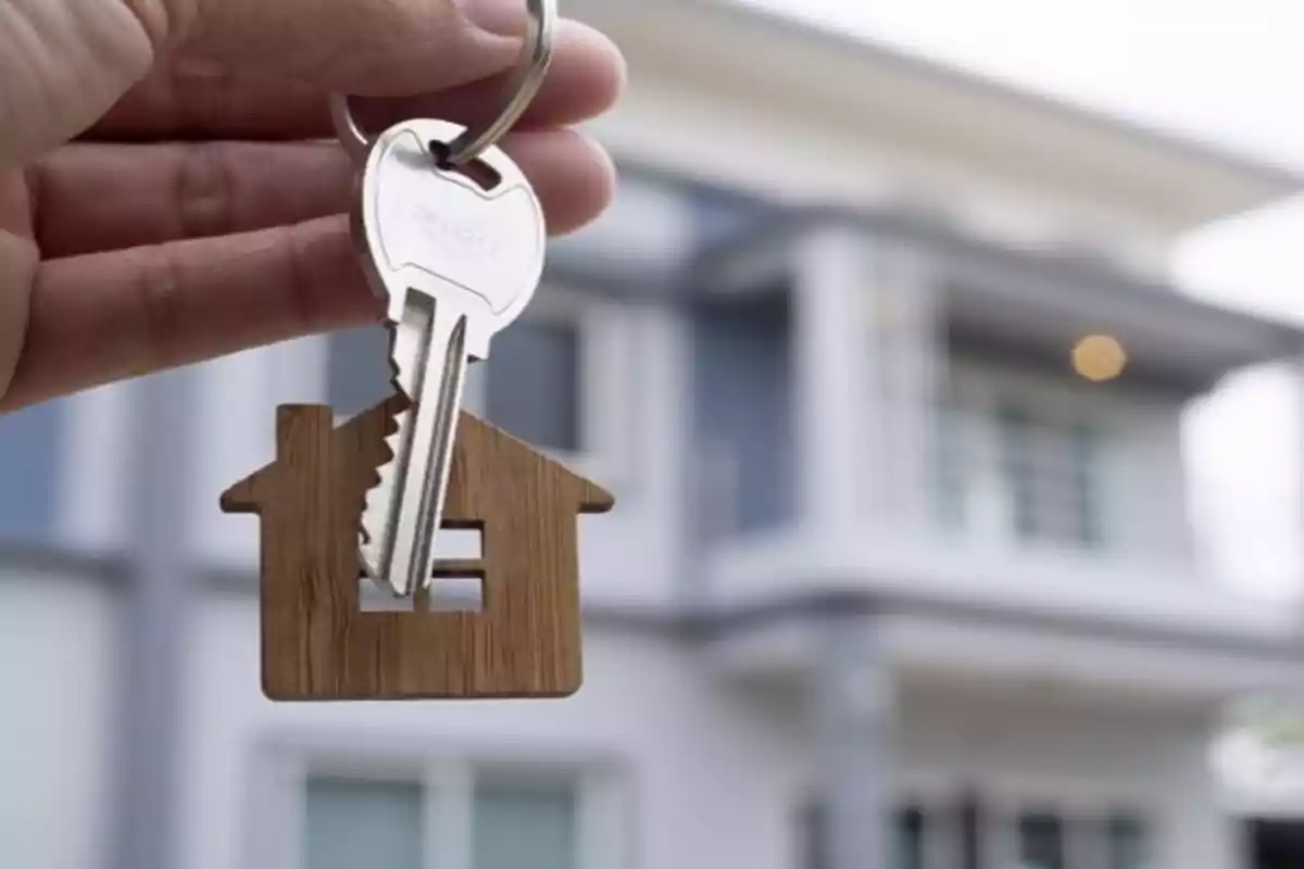 Una mano sosteniendo una llave con un llavero en forma de casa frente a una vivienda moderna. Una mano sosteniendo una llave con un llavero en forma de casa frente a una vivienda moderna.
