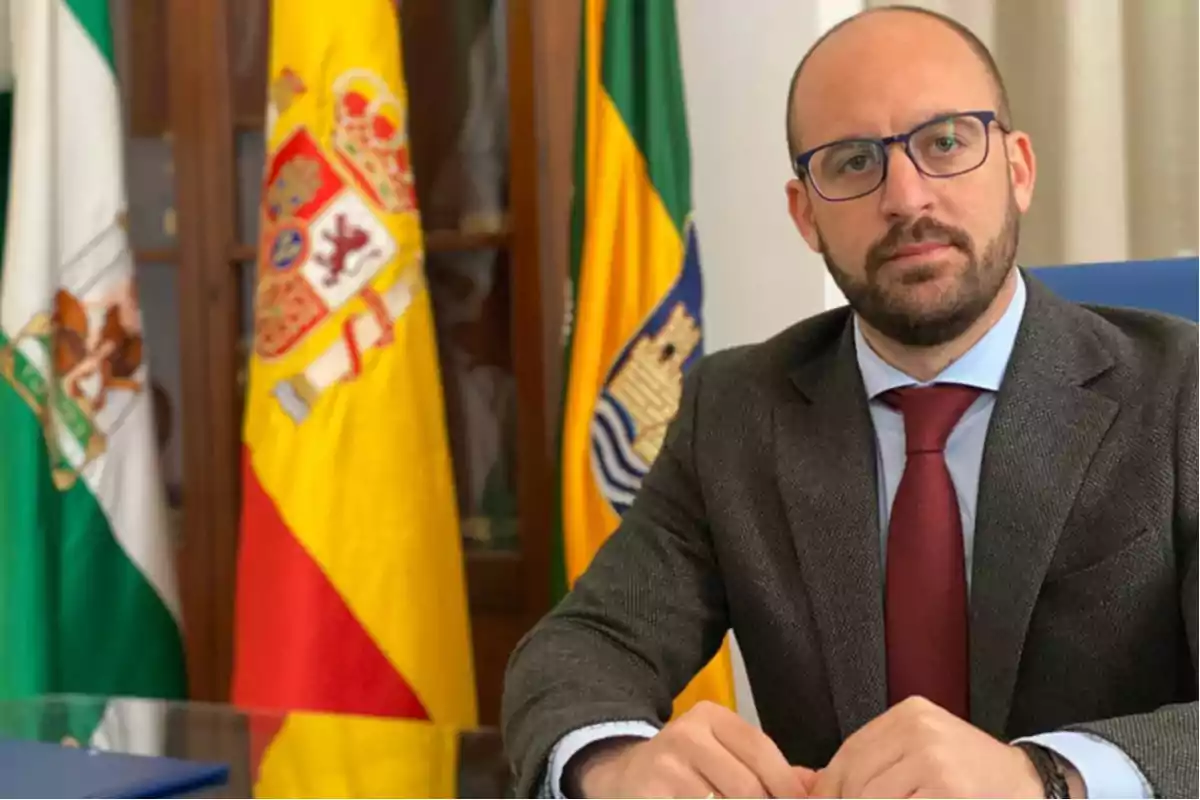 Un hombre con gafas y traje oscuro está sentado frente a banderas de España y Andalucía.