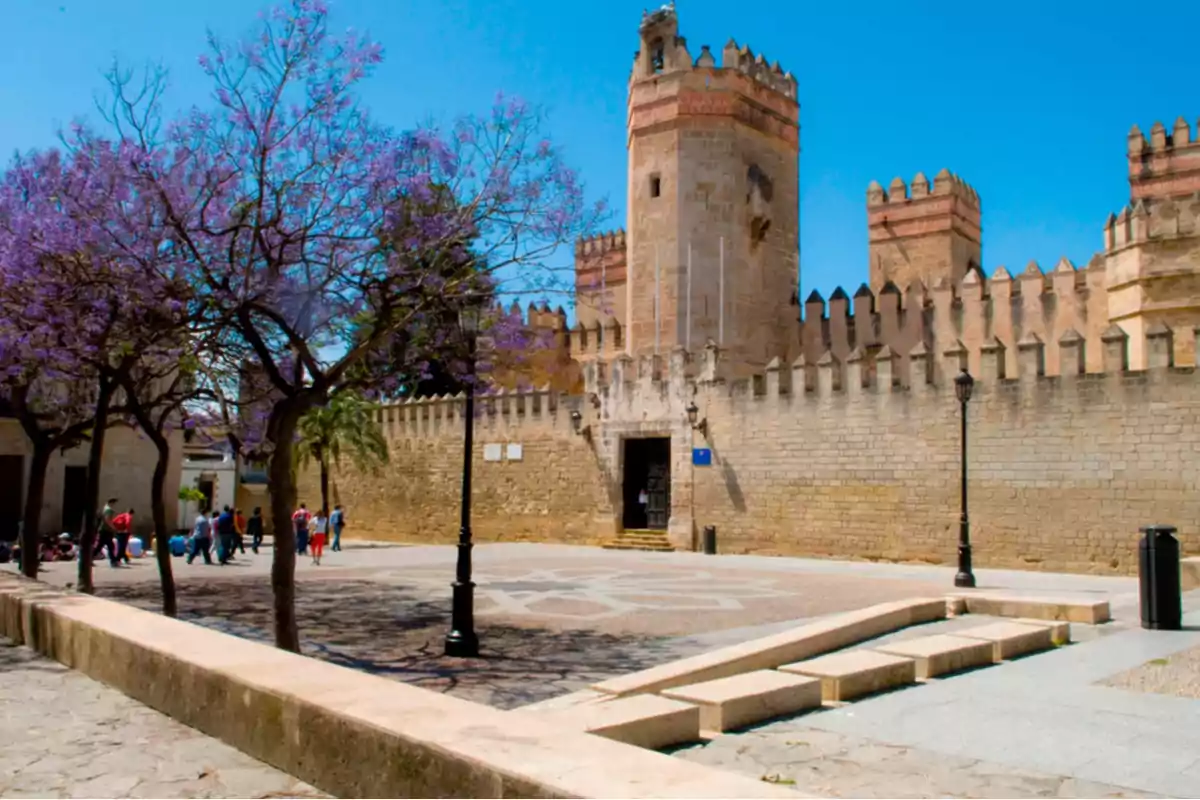 Vista de un castillo medieval con torres y murallas rodeado de árboles con flores moradas y un grupo de personas paseando en la plaza.