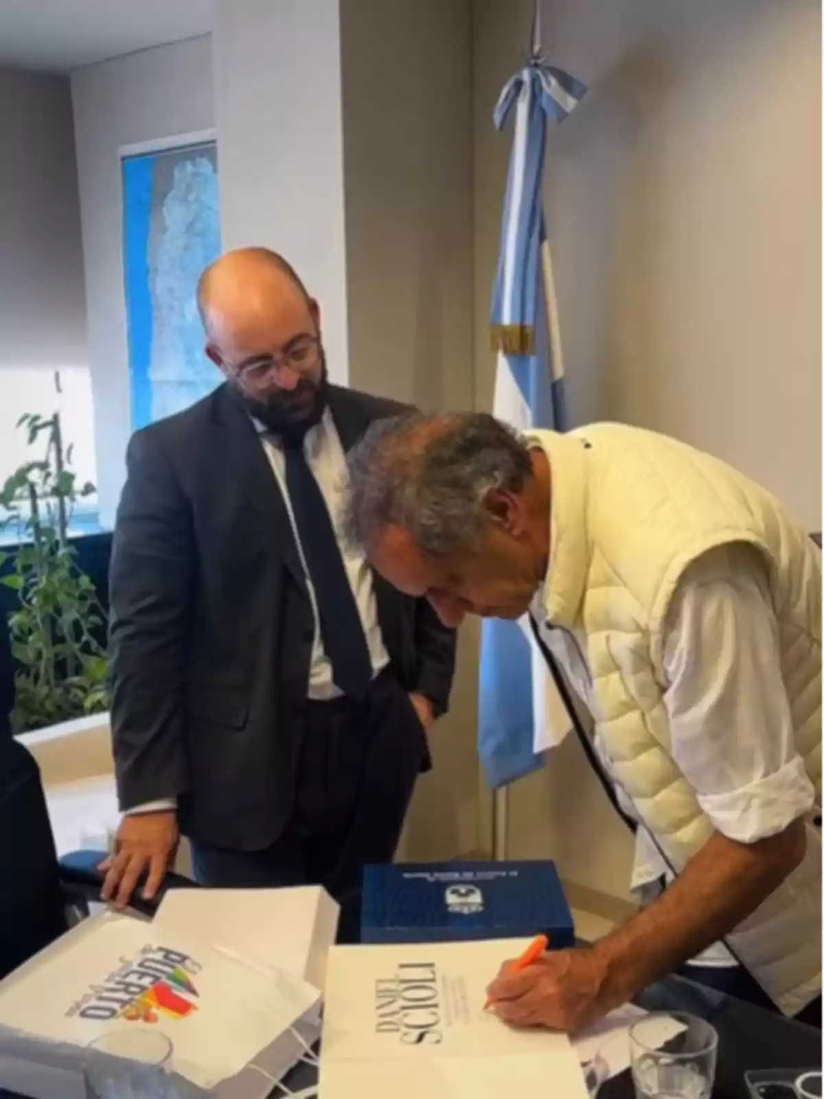 Dos personas están en una oficina; una de ellas está firmando un documento sobre una mesa mientras la otra observa. En el fondo se ve una bandera y un mapa.