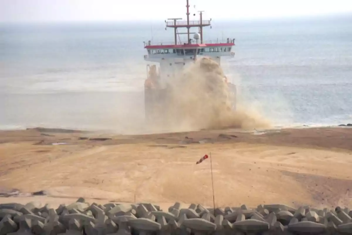 Un barco realiza trabajos de dragado en la costa, expulsando arena y agua hacia la orilla, con un rompeolas de bloques de concreto en primer plano.