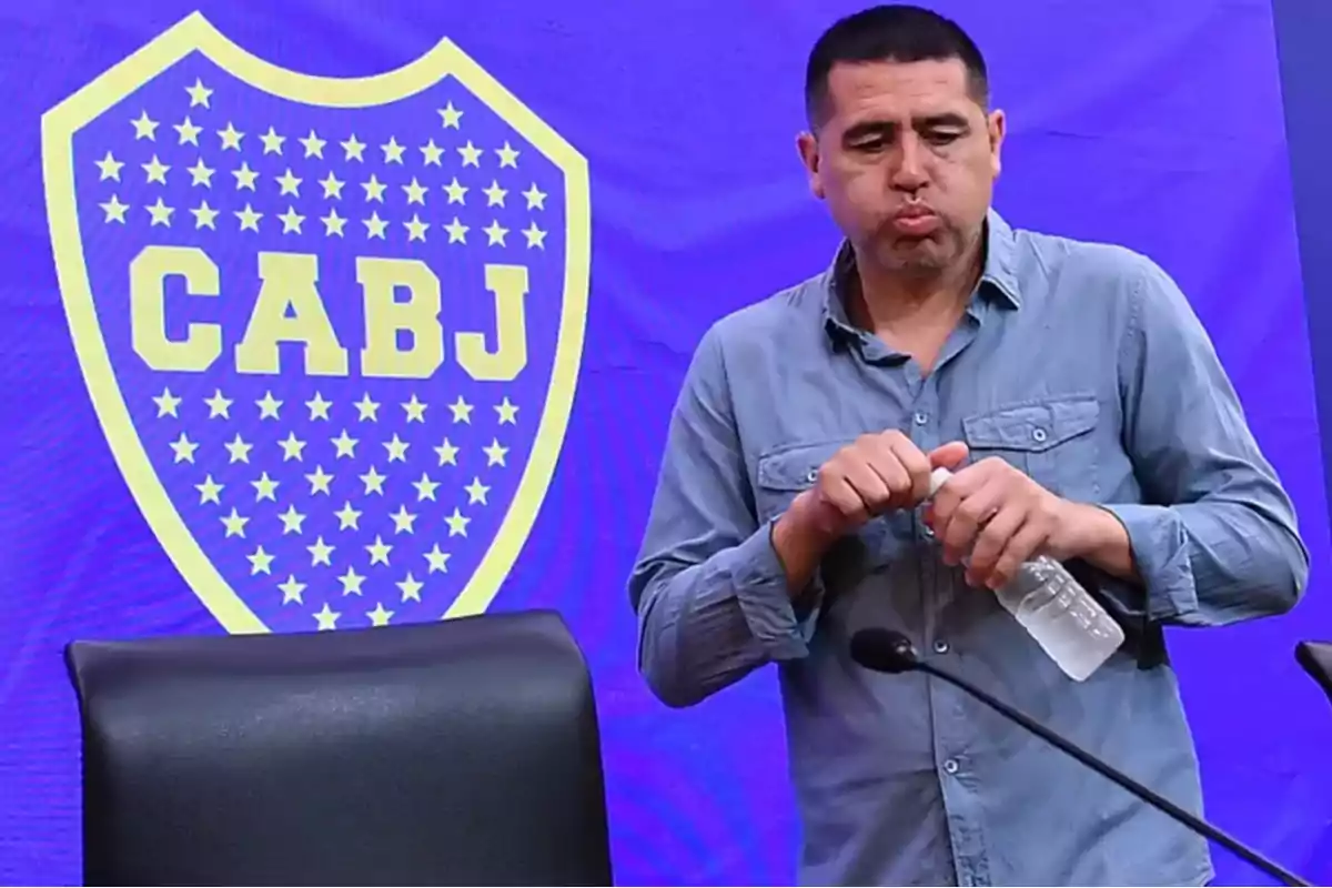 Hombre con camisa azul sosteniendo una botella de agua frente a un escudo de CABJ.