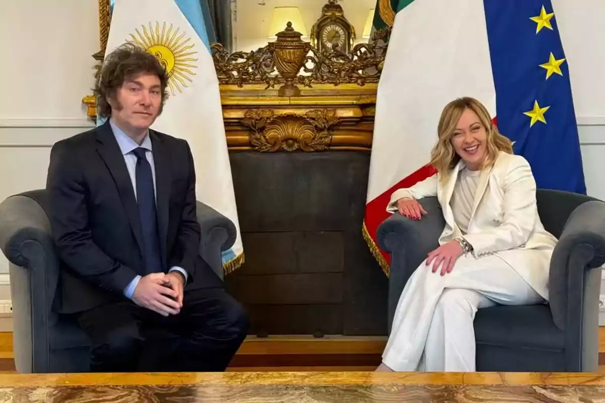 Dos personas sentadas en sillones frente a una chimenea con banderas de Argentina e Italia detrás.