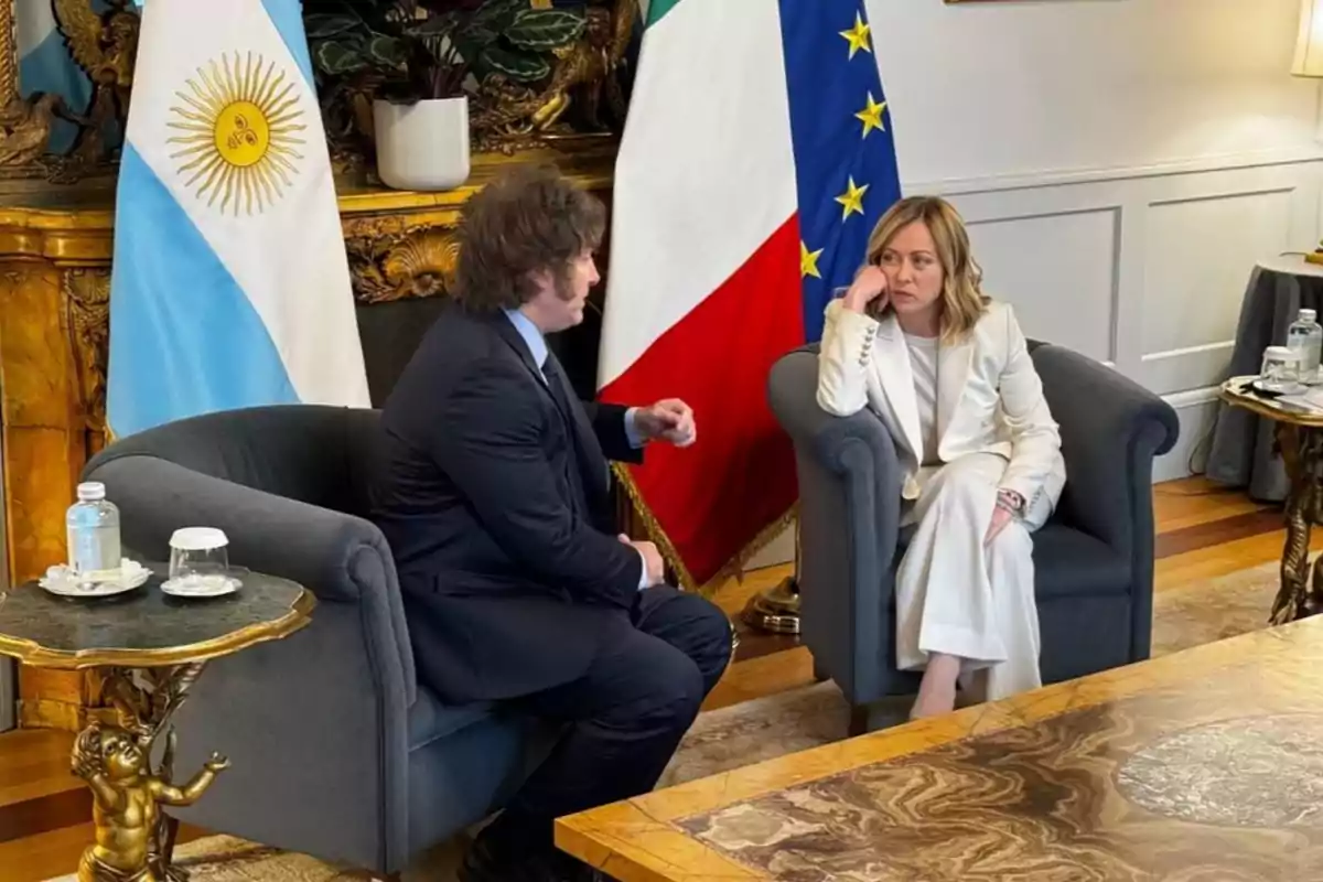 Dos personas sentadas en sillones conversan en una sala elegante con banderas de Argentina, Italia y la Unión Europea de fondo.