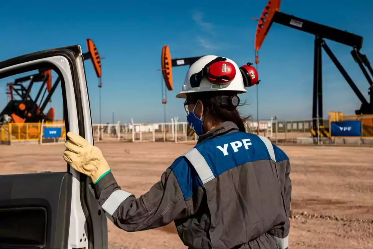 Trabajadora de YPF con casco y equipo de protección en un campo petrolero con bombas de extracción al fondo.