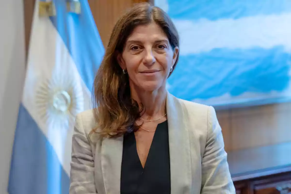 Una mujer de cabello castaño y chaqueta clara está de pie frente a una bandera argentina.