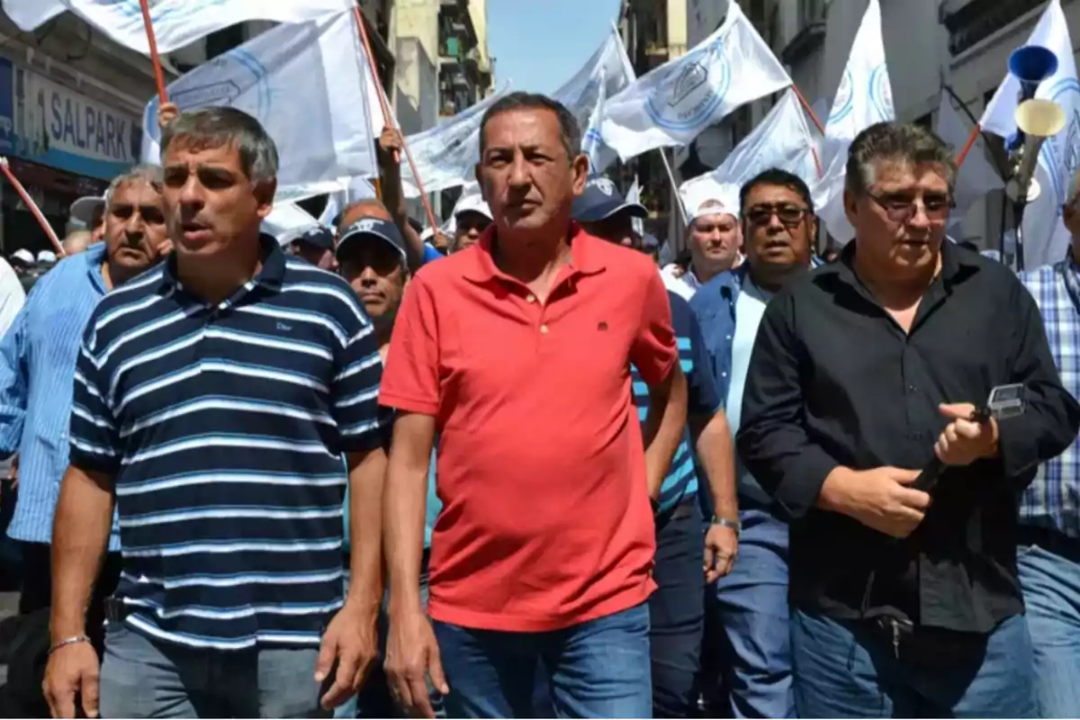 Un grupo de personas marcha por una calle con banderas blancas en el fondo.