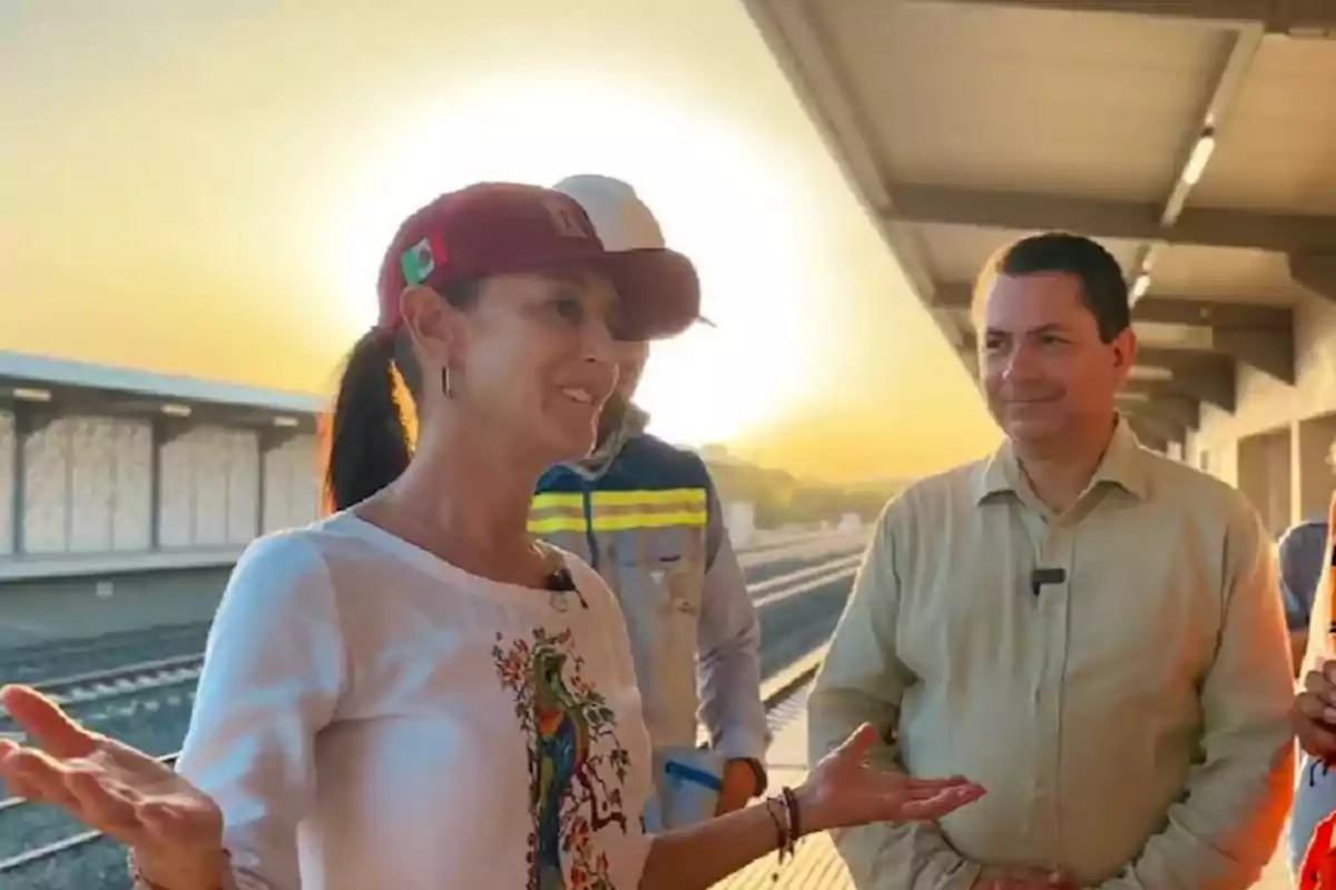 Una mujer con gorra roja y camiseta blanca está conversando con un grupo de personas en una estación de tren al atardecer.