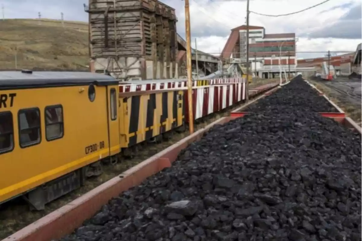 Un tren amarillo transporta carbón en una mina con edificios industriales al fondo.