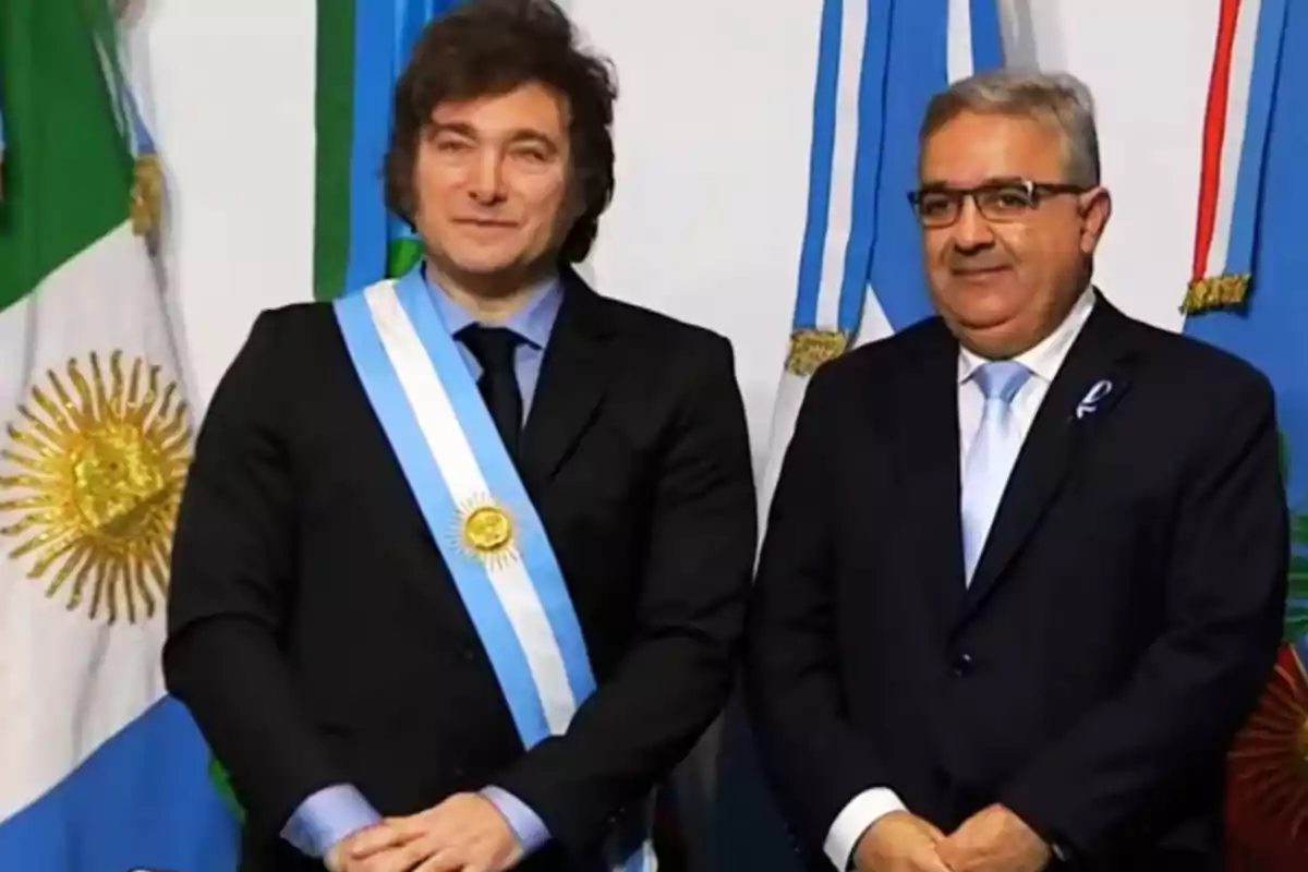 Dos hombres de pie con trajes formales y bandas presidenciales frente a banderas argentinas.