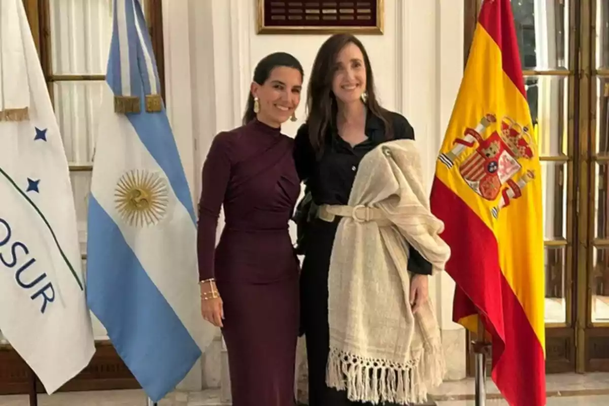 Dos mujeres posan juntas en un interior elegante con las banderas de Argentina y España a su lado.