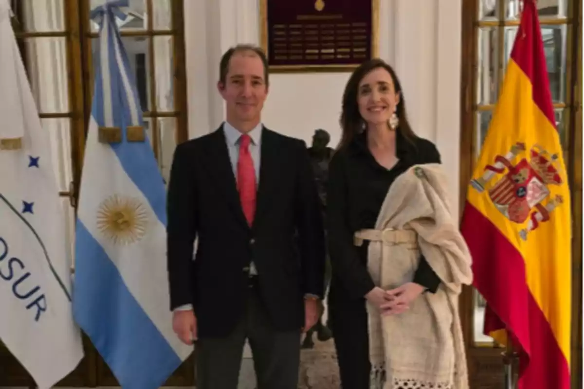 Dos personas posan juntas en un interior elegante, flanqueadas por las banderas de Argentina y España.