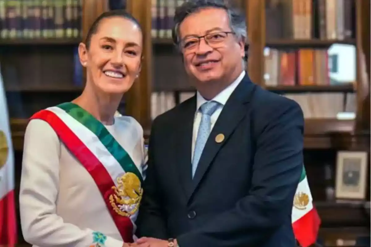 Dos personas posan juntas en un entorno formal con banderas y estanterías de libros al fondo.