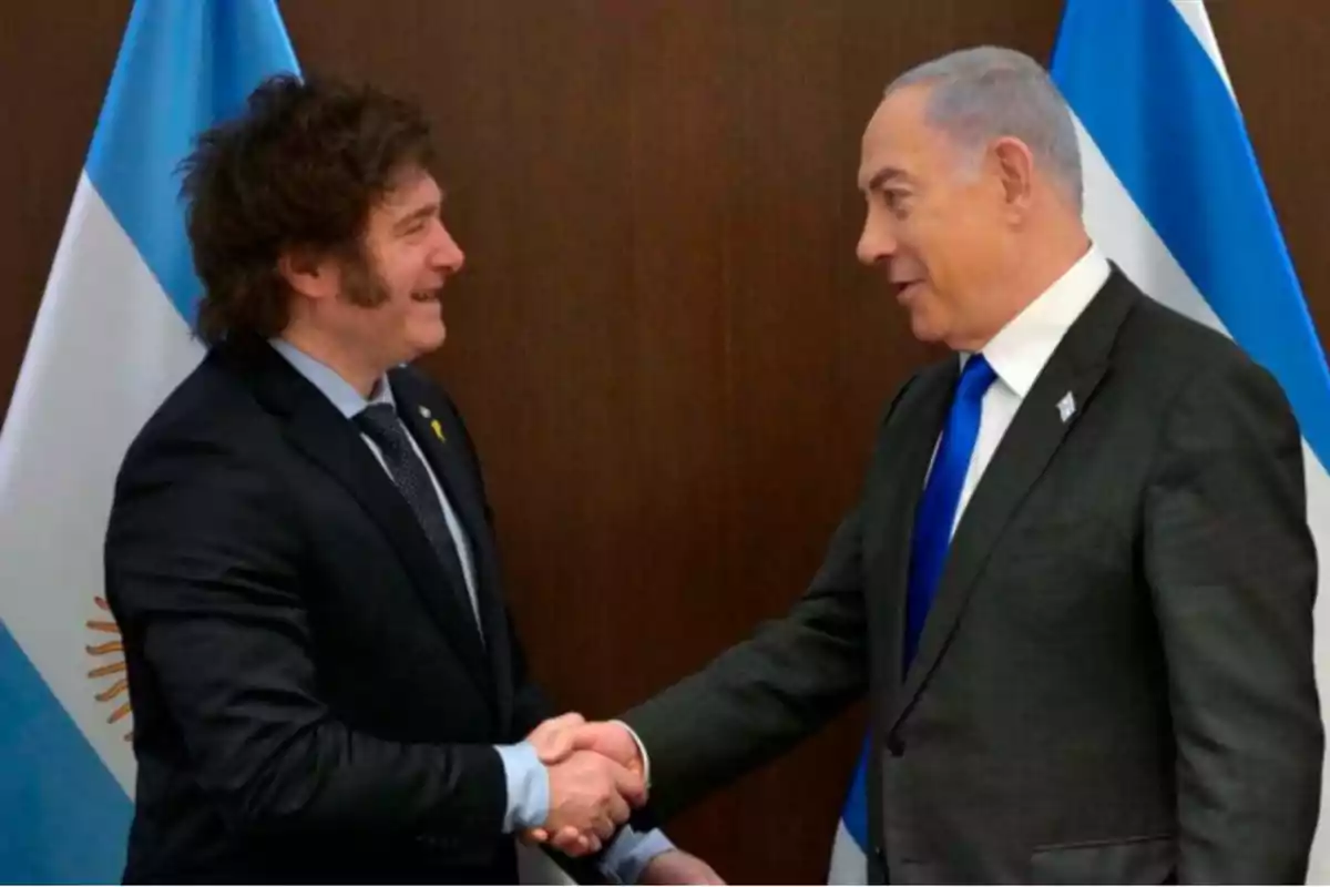 Dos hombres de traje se dan la mano frente a banderas de Argentina e Israel.