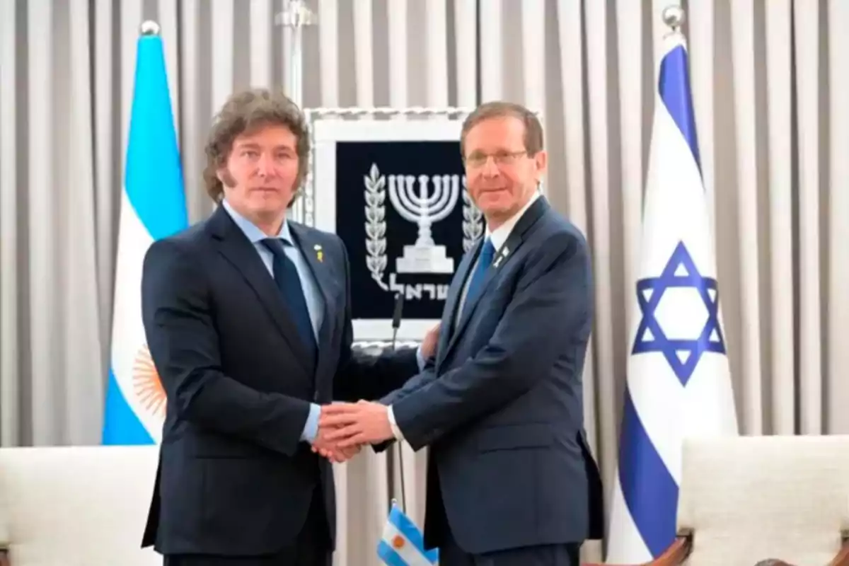 Dos hombres de traje se dan la mano frente a las banderas de Argentina e Israel.