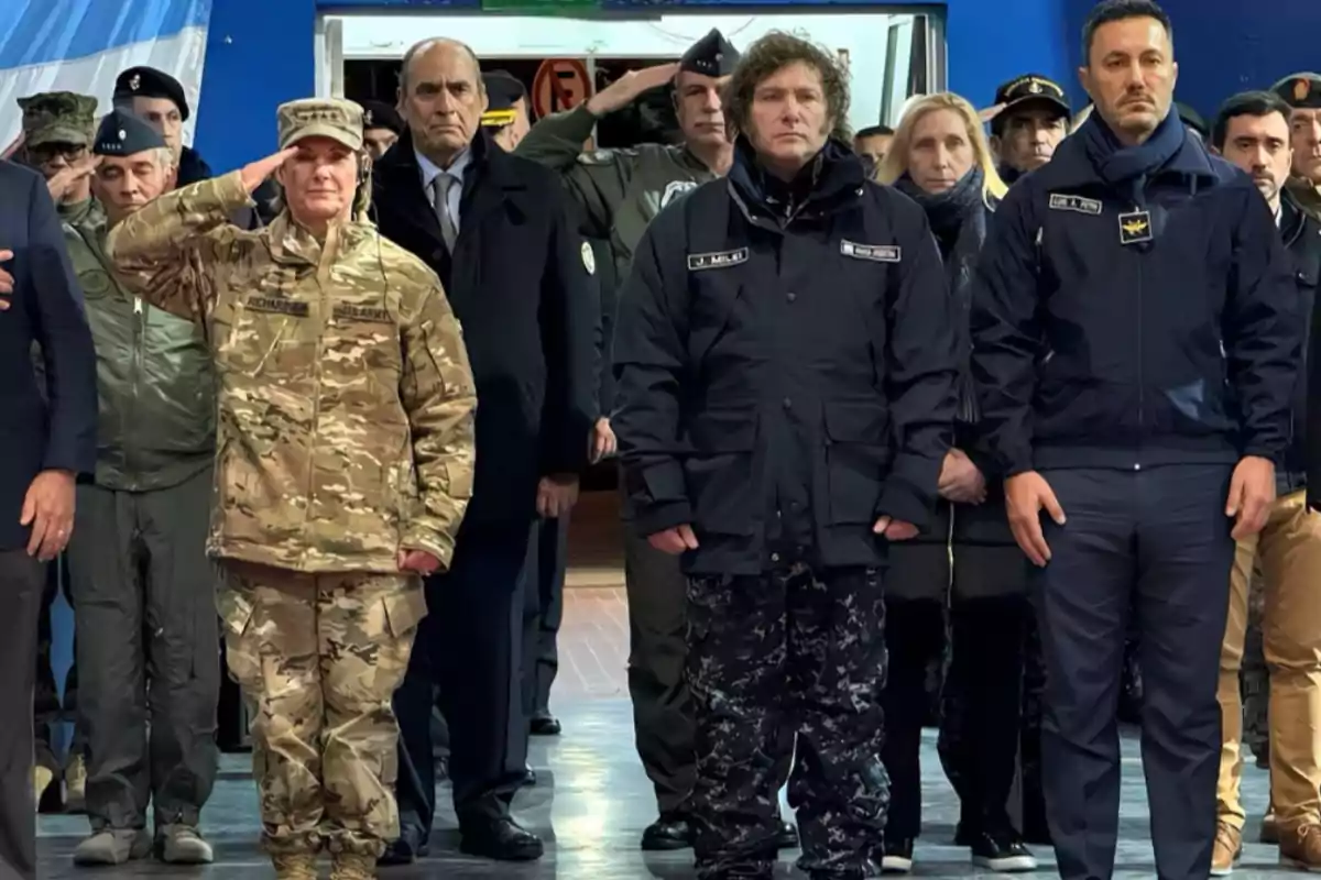 Un grupo de personas en uniforme militar y civil se encuentra de pie en una ceremonia solemne, con una bandera en el fondo.