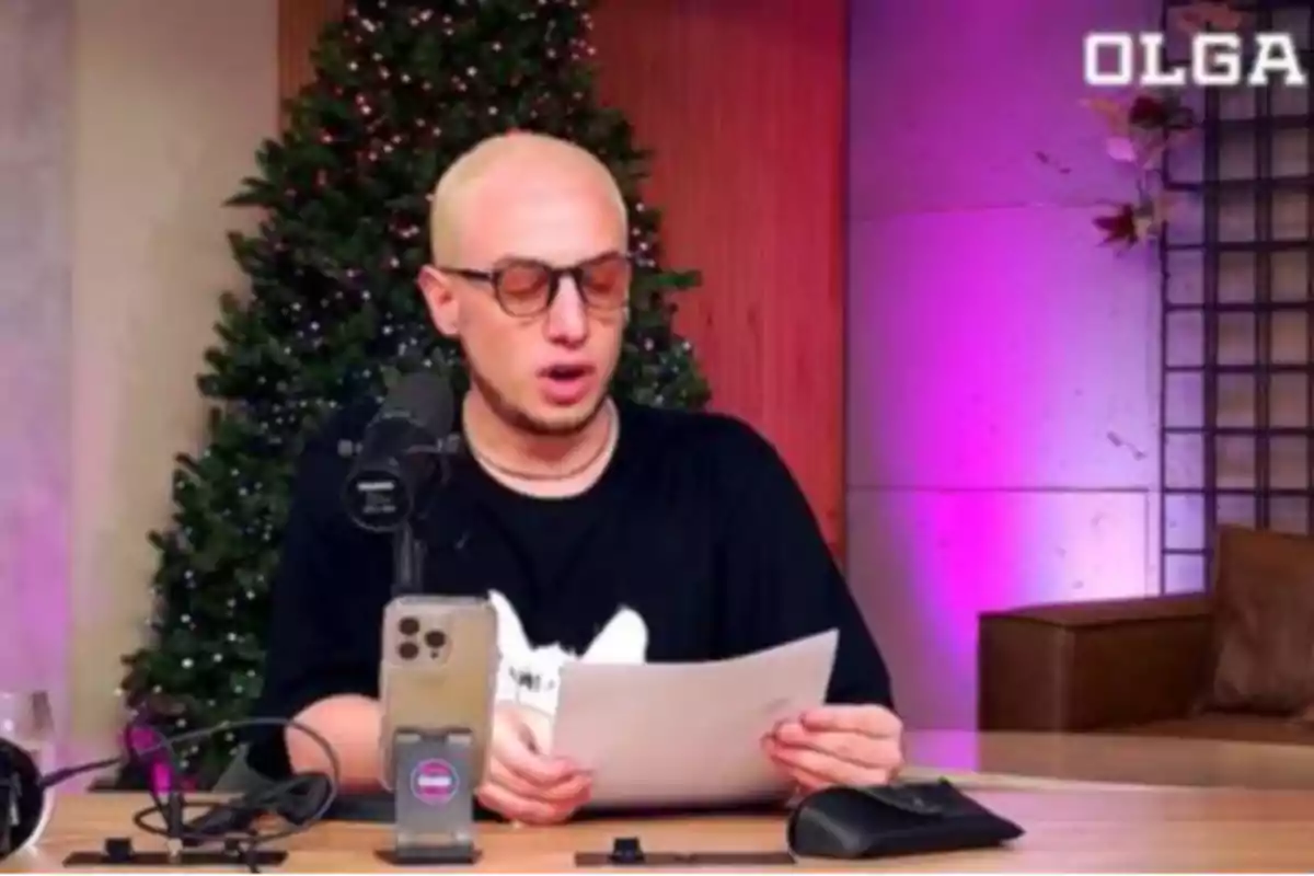 Una persona con gafas y cabello rubio está sentada frente a un micrófono y sostiene un papel, con un árbol de Navidad decorado al fondo y luces de color púrpura en la pared.