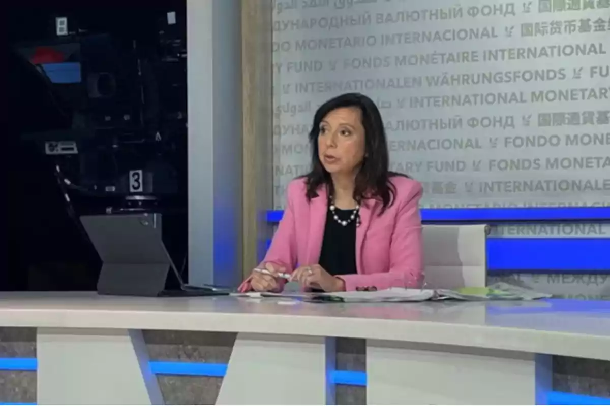 Una mujer con chaqueta rosa está sentada en un estudio de televisión frente a una mesa con papeles y una computadora portátil, mientras una cámara de televisión está en el fondo.