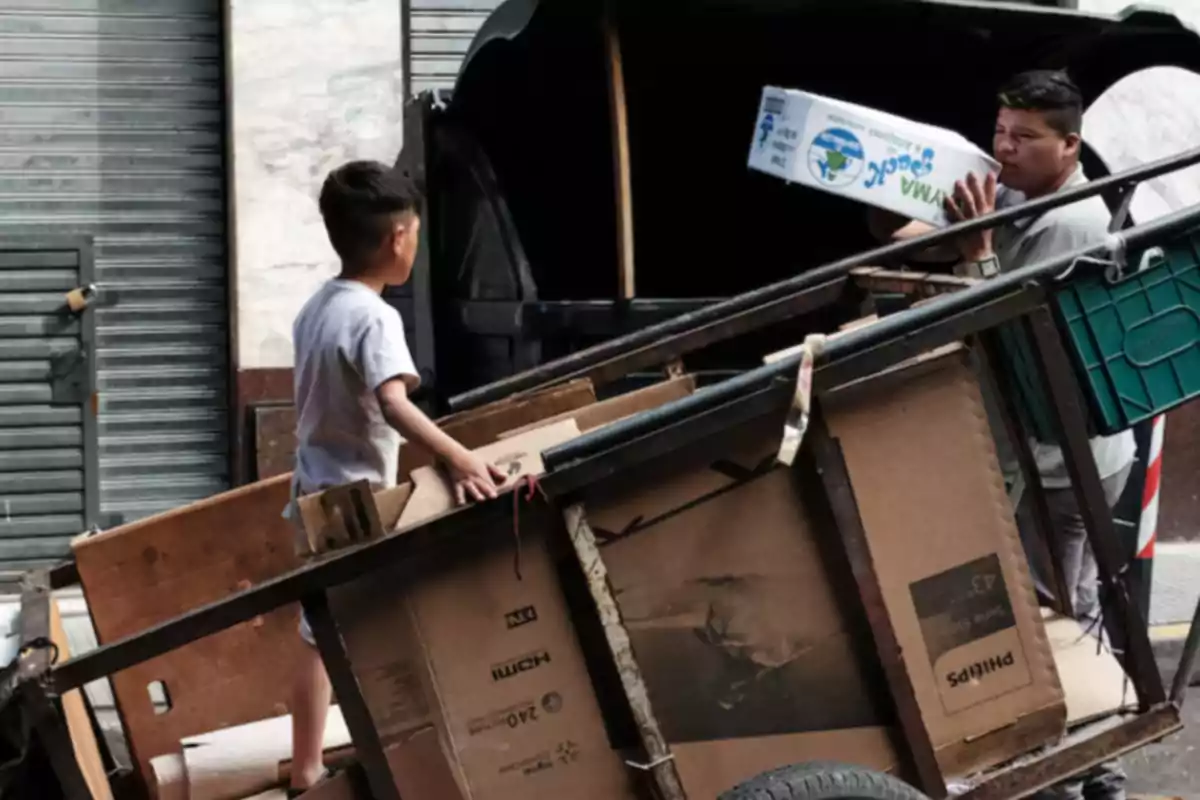 Un niño y un hombre cargan una carreta con cajas de cartón en una calle urbana.