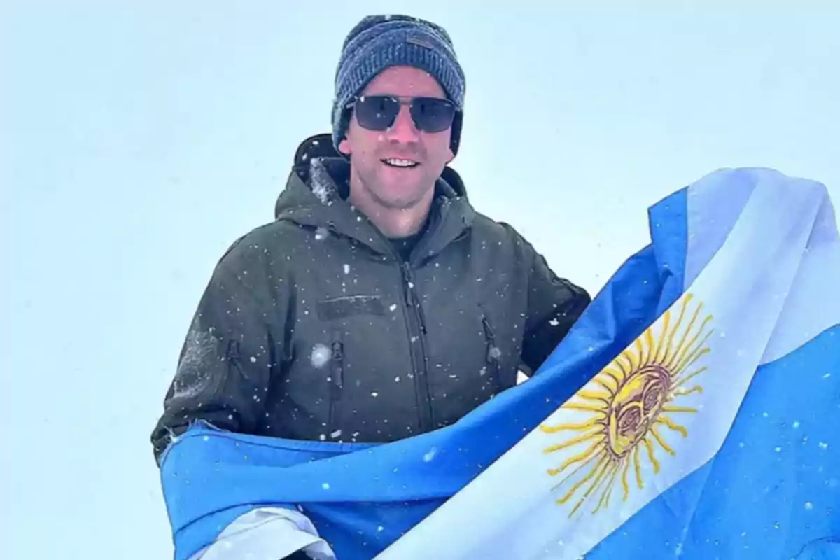 Persona con gafas de sol y gorro sosteniendo una bandera argentina en un entorno nevado.