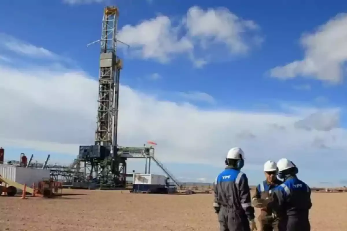 Tres trabajadores con cascos y uniformes de YPF conversan cerca de una torre de perforación en un campo petrolero bajo un cielo azul con nubes.