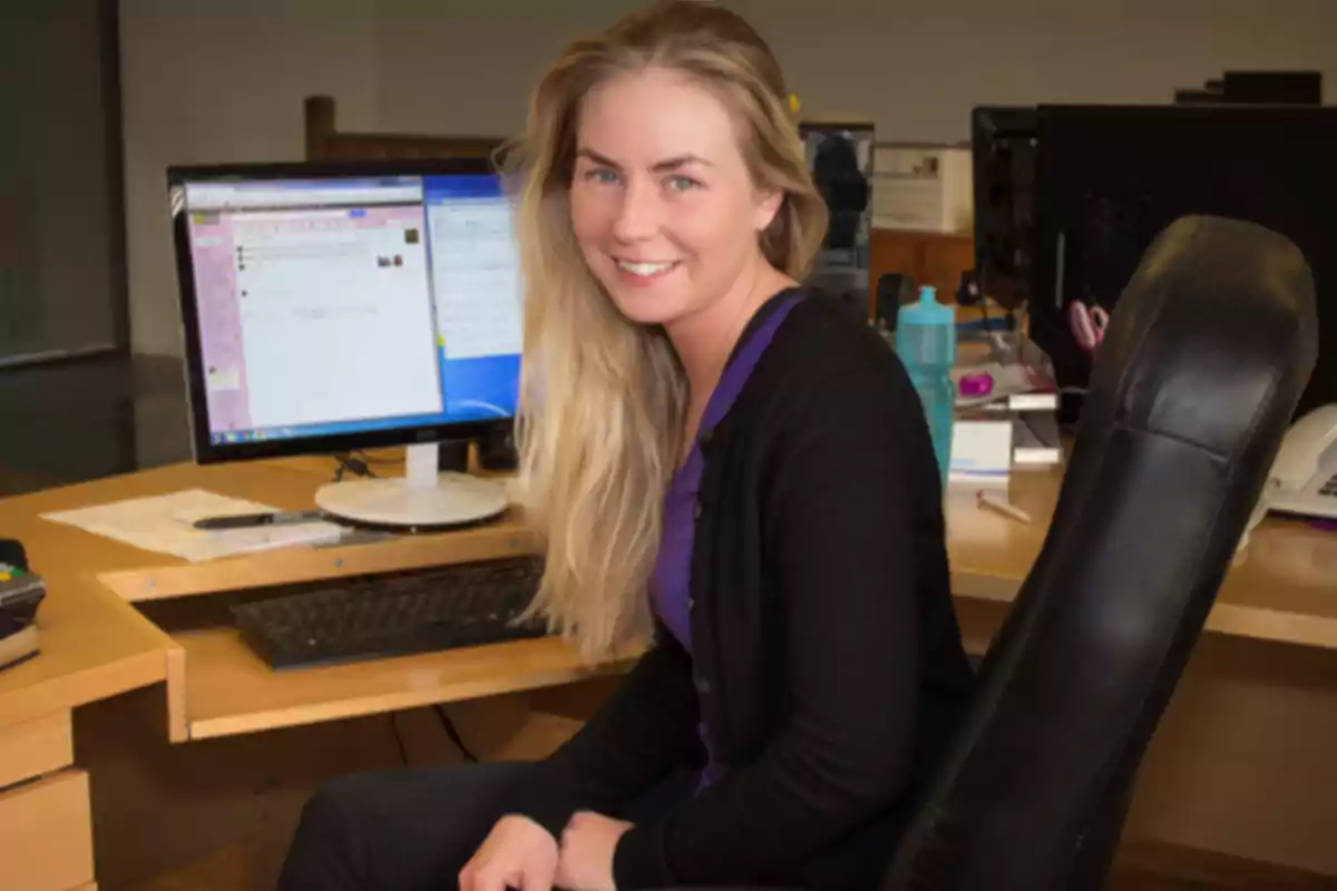 Una mujer sonriente sentada en una silla de oficina frente a un escritorio con una computadora y varios objetos.