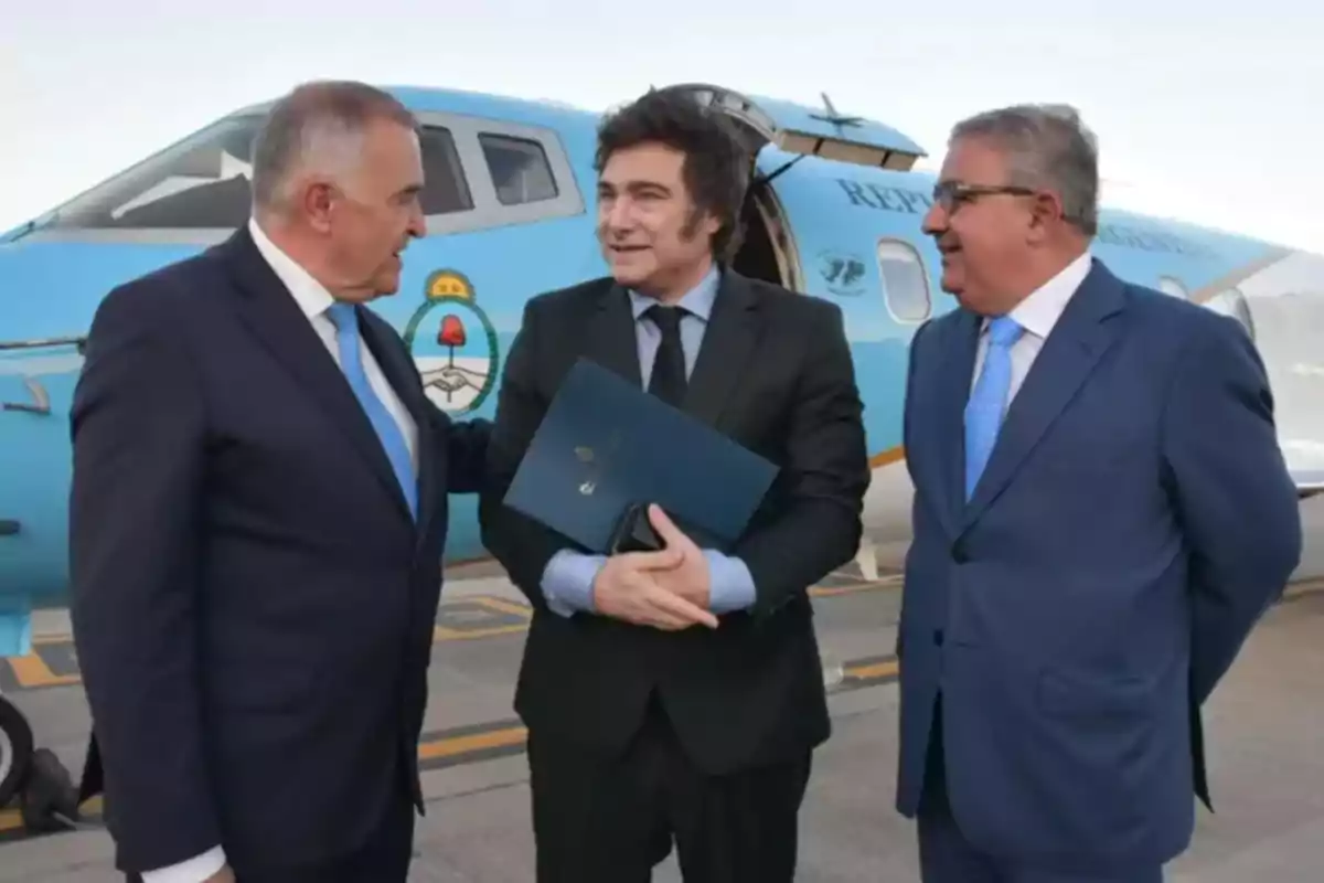 Tres hombres de traje conversan frente a un avión azul con el escudo de Argentina.