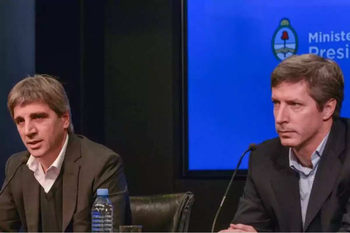 Dos hombres en una conferencia de prensa con un fondo azul que muestra un logotipo gubernamental.