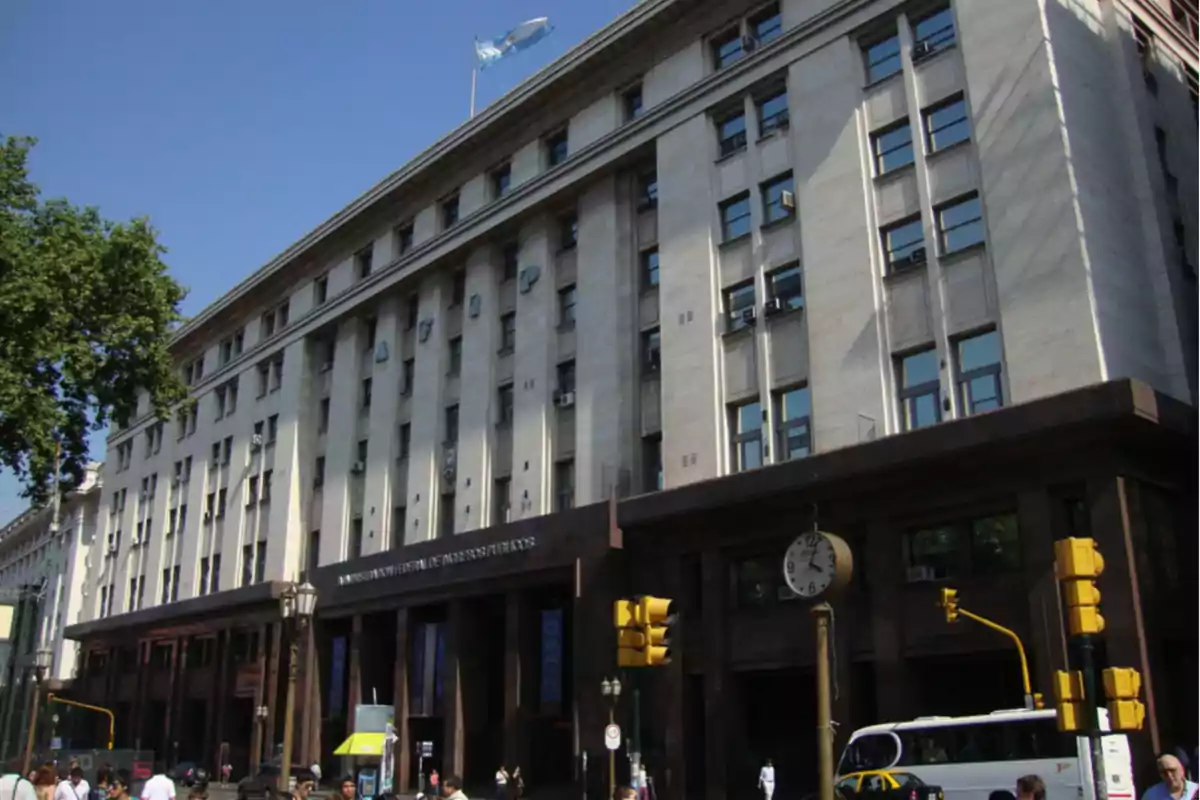 Edificio gubernamental con fachada de piedra y ventanas rectangulares en un día soleado con una bandera ondeando en la parte superior.