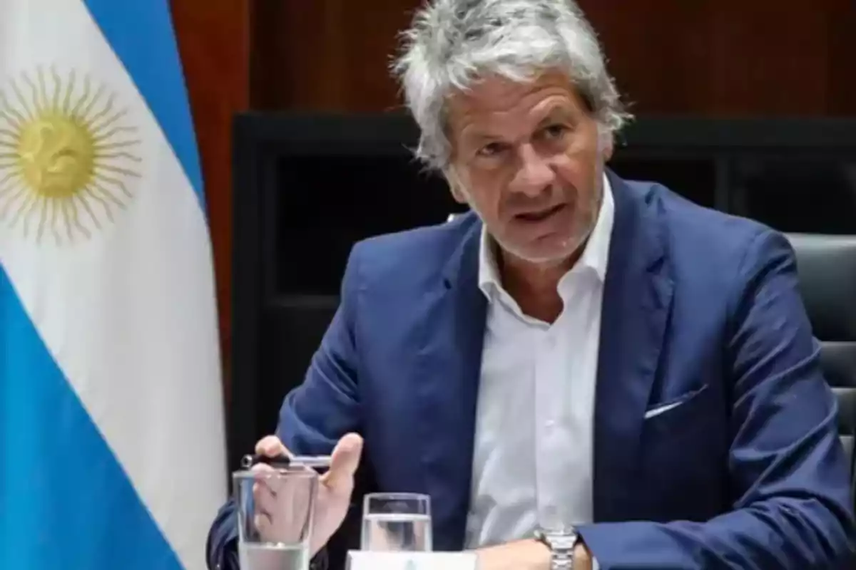 Hombre de cabello canoso en traje azul sentado en una mesa con un vaso de agua y una bandera de Argentina al fondo.