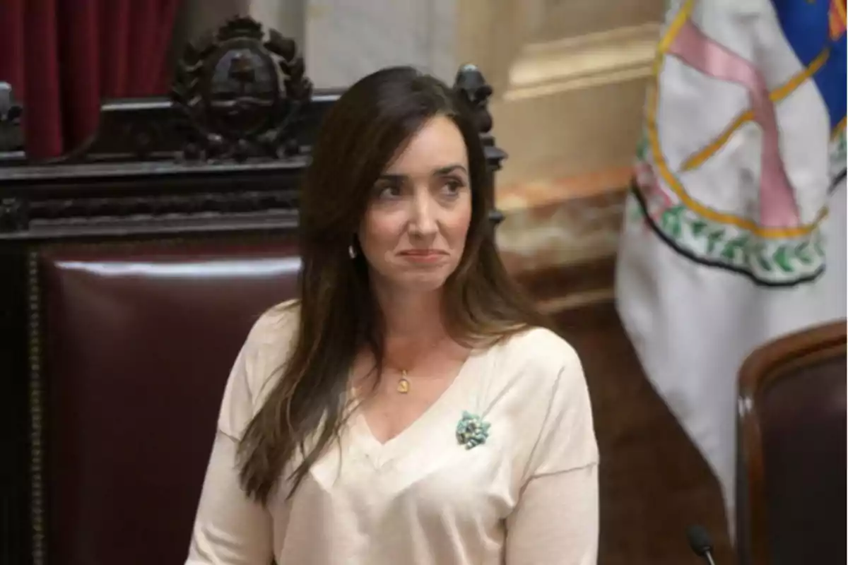 Una mujer con cabello largo y oscuro está sentada en una silla de madera con un respaldo decorativo y una bandera en el fondo.