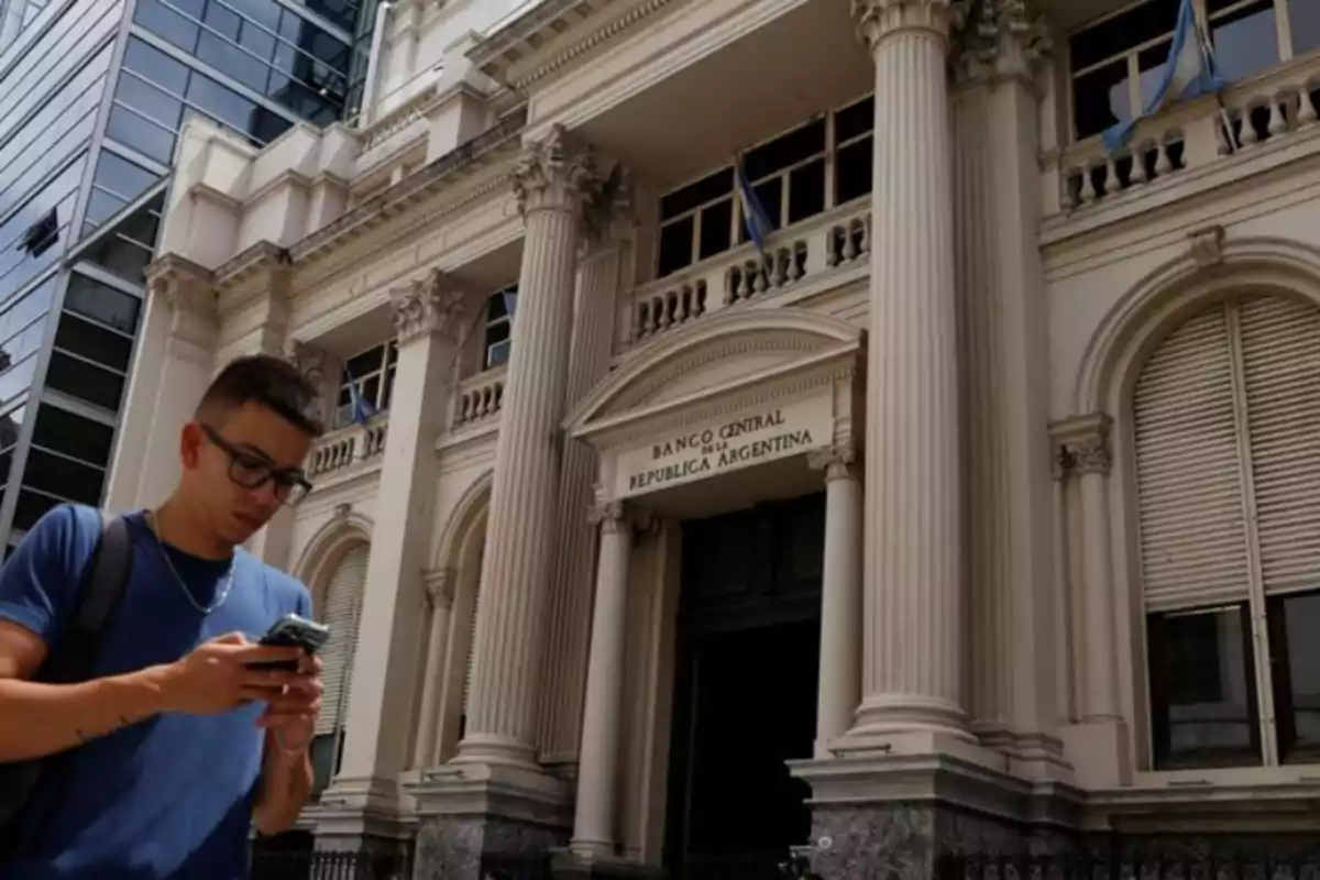 Un hombre con gafas y mochila revisa su teléfono frente a un edificio con columnas clásicas y un cartel que dice Un hombre con gafas y mochila revisa su teléfono frente a un edificio con columnas clásicas y un cartel que dice