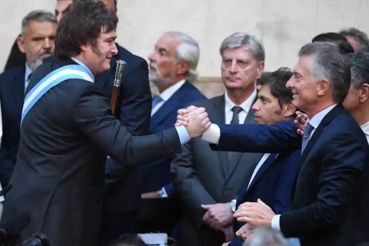 Dos hombres de traje se saludan con un apretón de manos en un evento formal rodeados de otras personas.