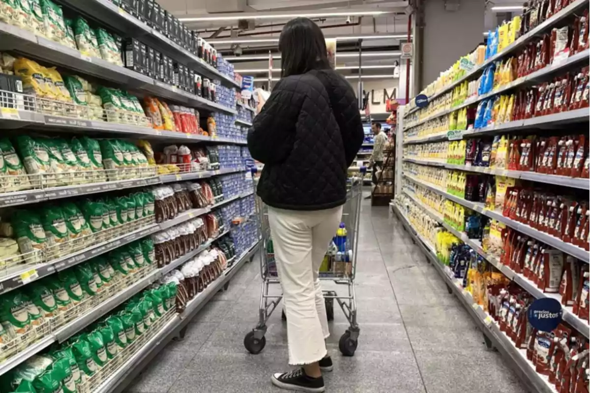 Una persona empuja un carrito de compras por un pasillo de supermercado lleno de productos en las estanterías.