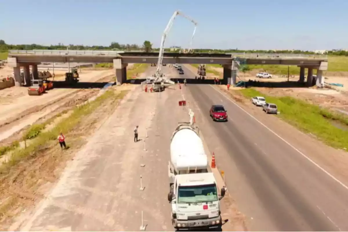 Camión de cemento en una carretera en construcción con un puente elevado y varios vehículos alrededor.