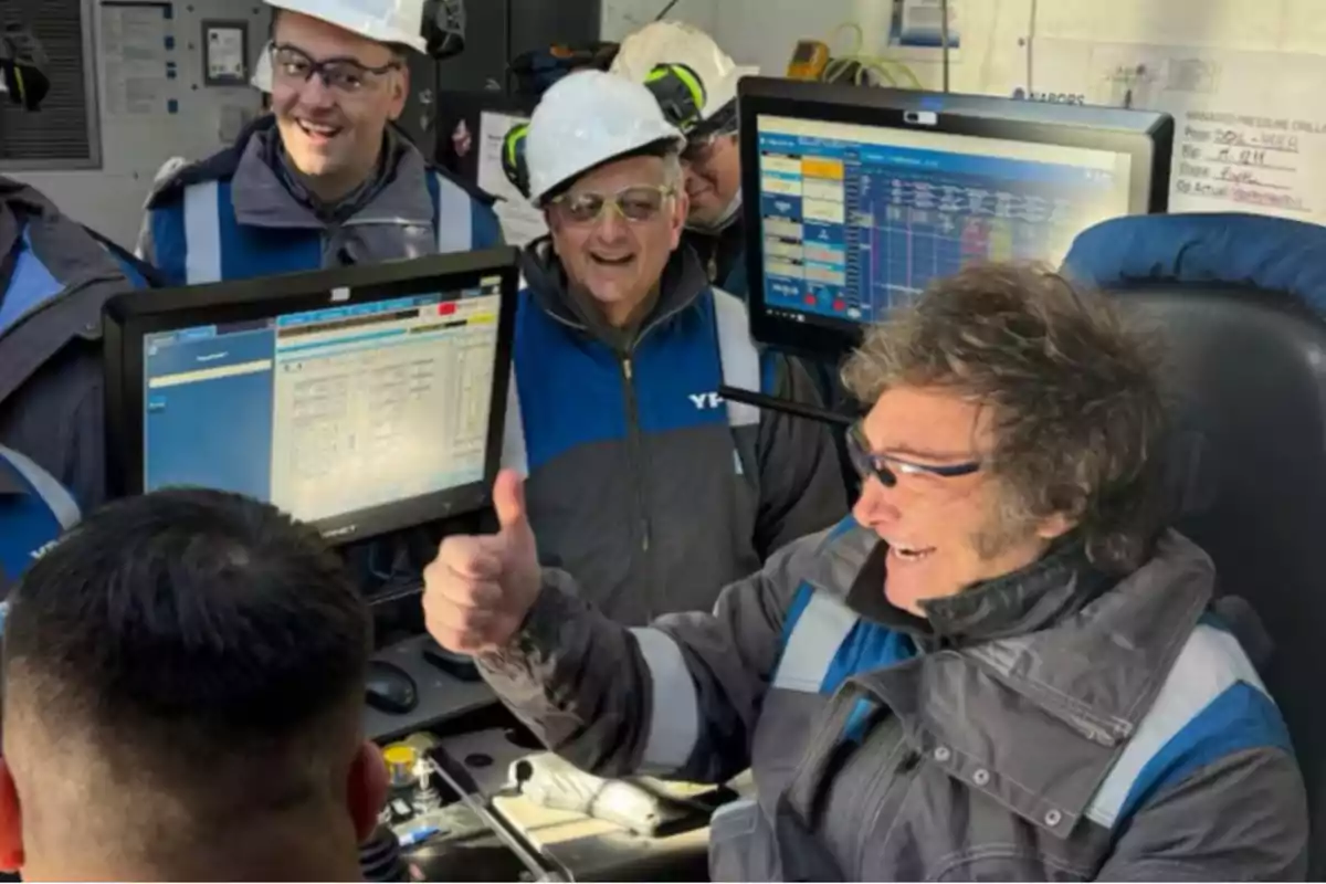 Un grupo de personas con cascos y gafas de seguridad sonríen mientras observan pantallas de computadora en un entorno industrial.