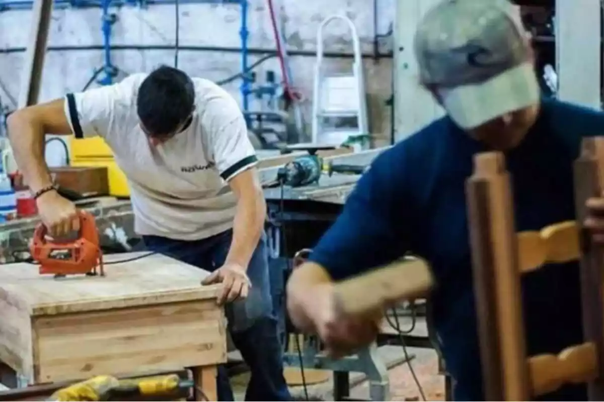 Dos personas trabajando en un taller de carpintería, una usando una sierra eléctrica y la otra ensamblando una estructura de madera.