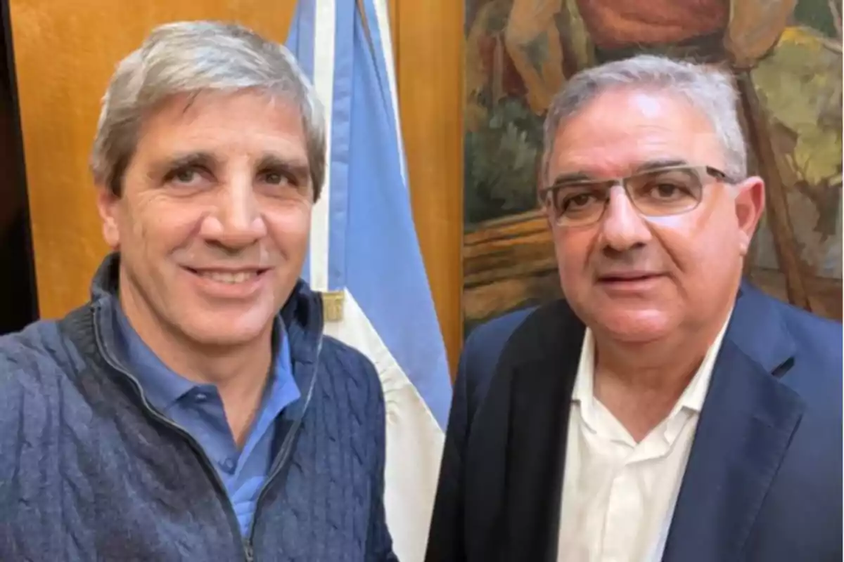 Dos hombres posan juntos frente a una bandera y un cuadro en un entorno interior.