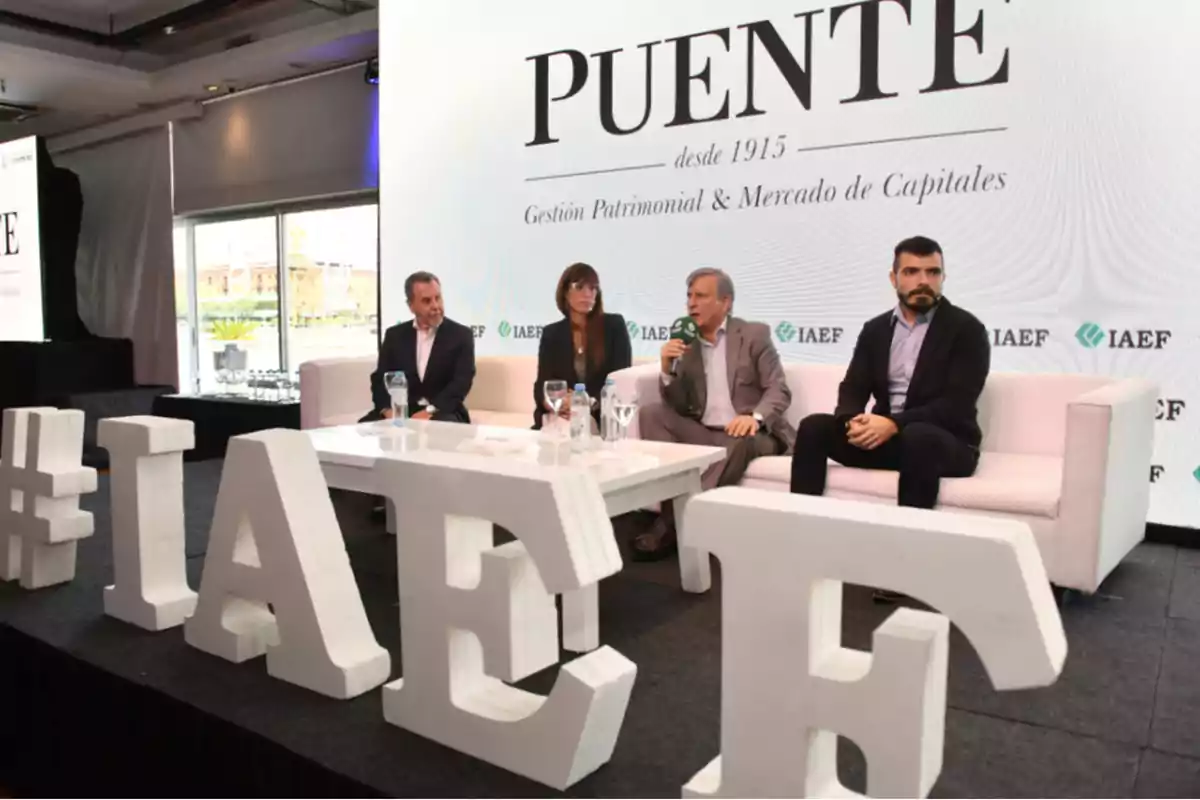 Un grupo de personas está sentado en un escenario durante un evento de Puente y IAEF, con letras grandes formando un hashtag en primer plano.