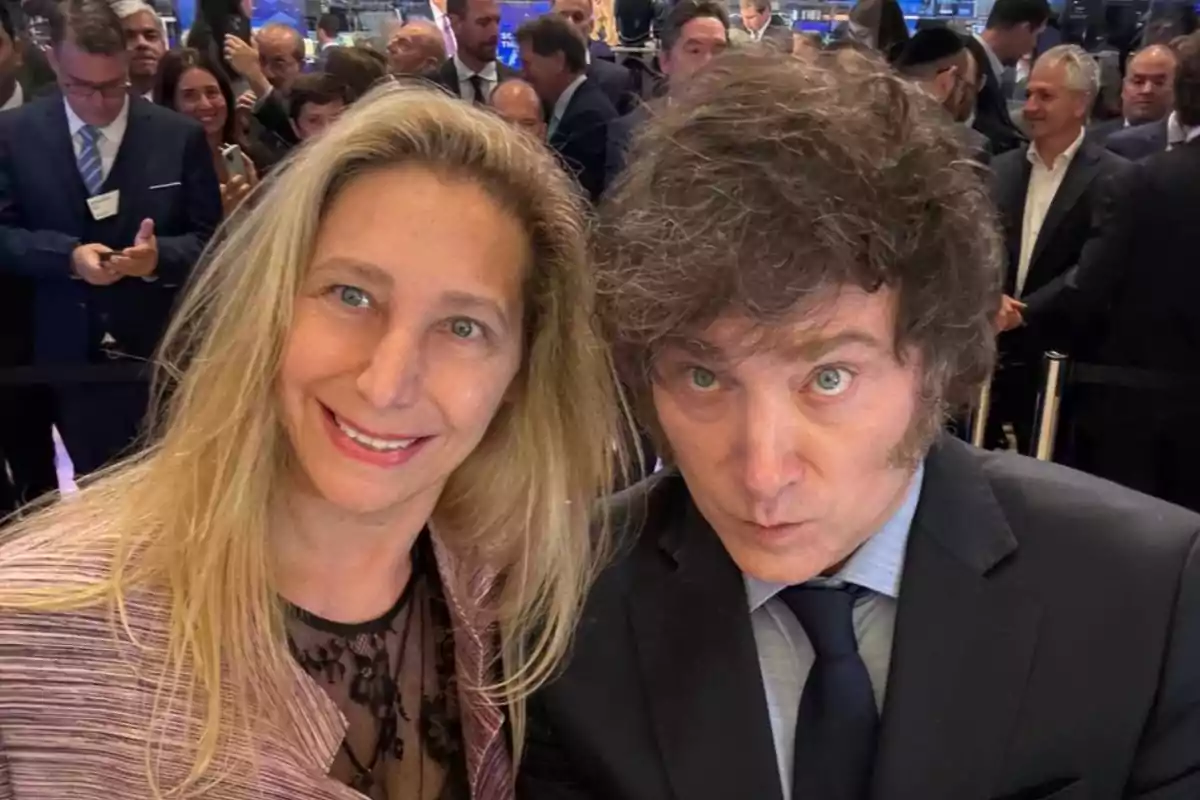 Dos personas posando para una selfie en un evento formal con varias personas al fondo.