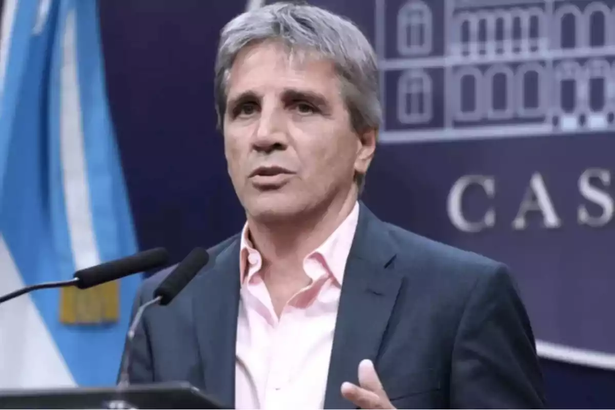 Un hombre de cabello canoso y traje oscuro habla frente a un micrófono con una bandera y un fondo azul detrás.