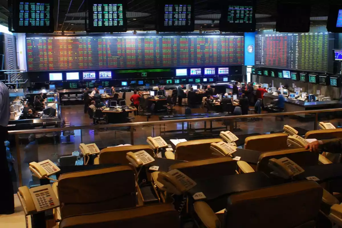 Sala de operaciones de una bolsa de valores con múltiples pantallas mostrando datos financieros y personas trabajando en escritorios.