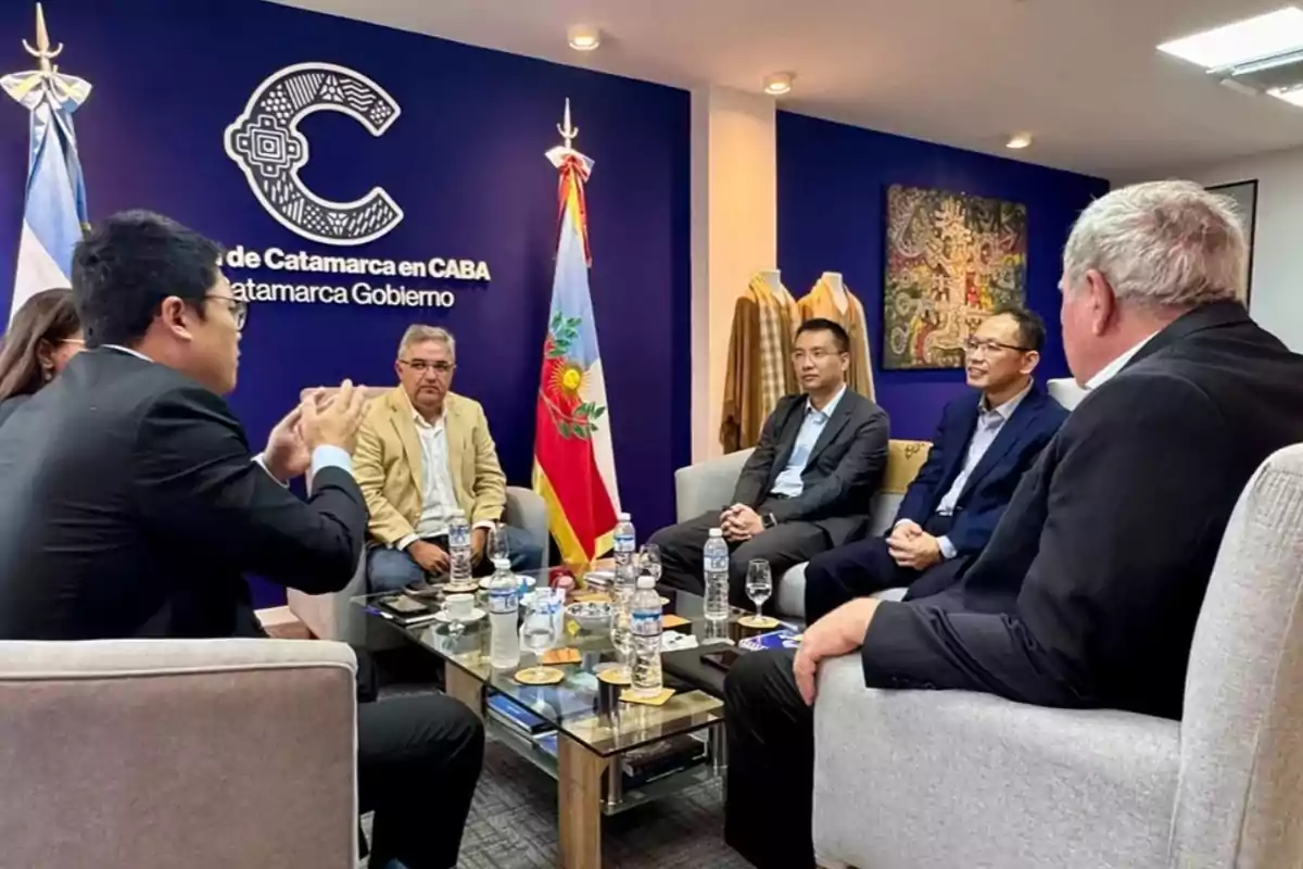 Un grupo de personas en una reunión formal sentadas alrededor de una mesa con botellas de agua y tazas, en una sala con banderas y un logotipo en la pared.