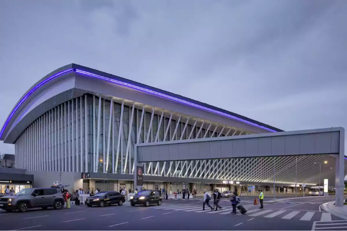 Edificio moderno con fachada de vidrio y estructura de soporte blanca, iluminado con luces azules en el techo, con personas y vehículos en movimiento en la calle frente a él.