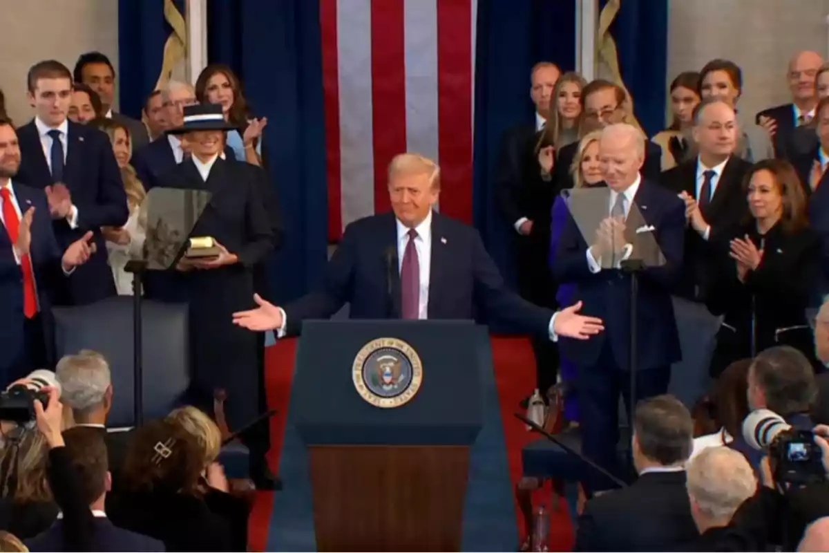 Un hombre de traje está en un podio con el sello presidencial, rodeado de personas que aplauden, con una bandera de fondo.