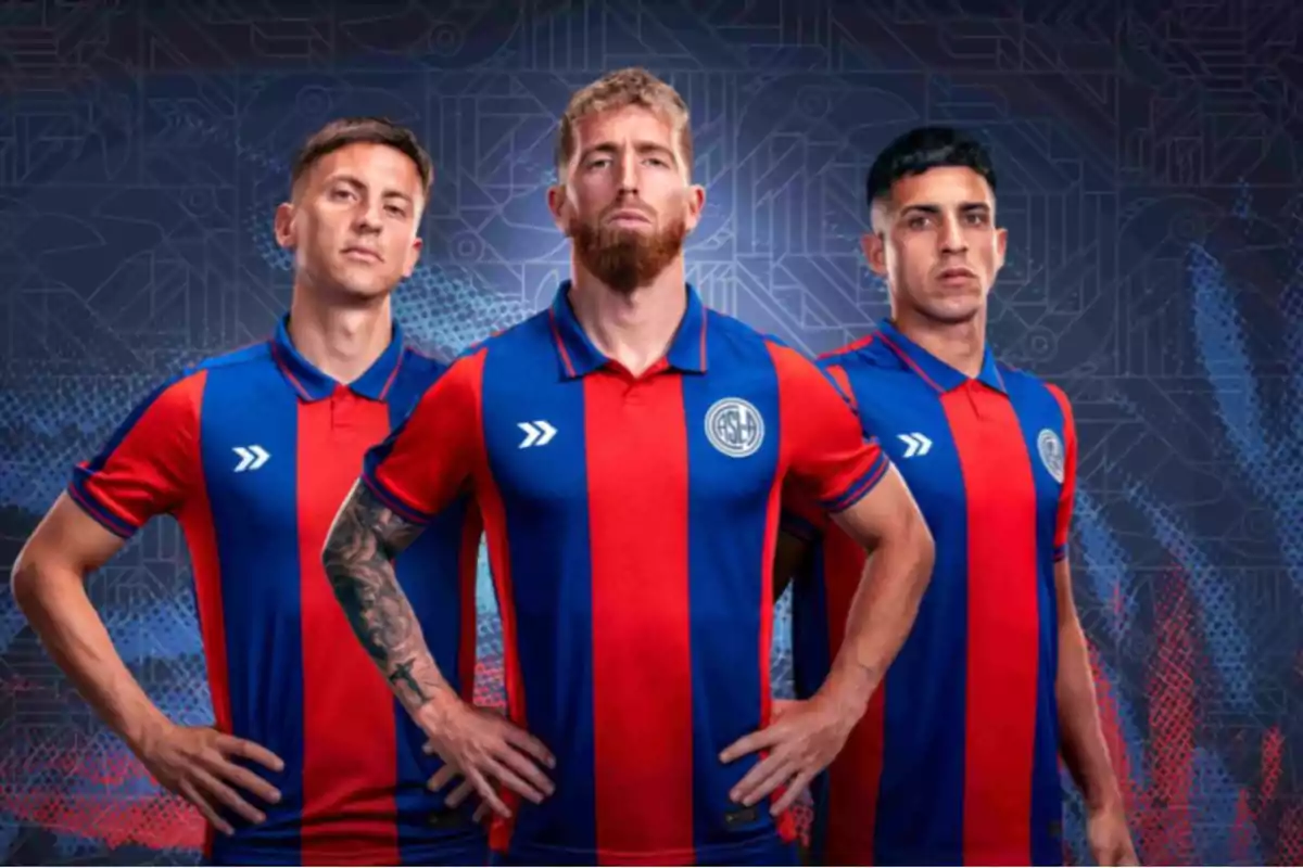 Tres jugadores de fútbol posan con uniformes azul y rojo frente a un fondo con diseño geométrico.