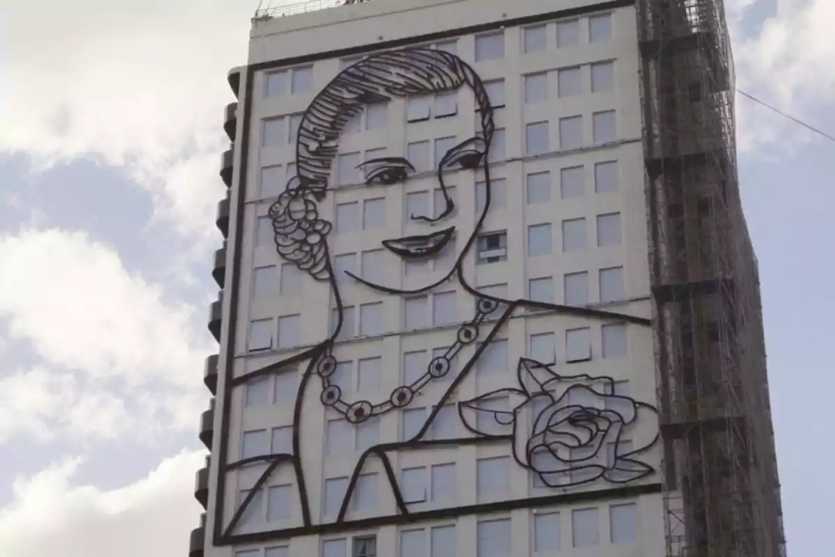 Un edificio alto con una gran imagen de una mujer dibujada en su fachada, con el cielo parcialmente nublado de fondo.