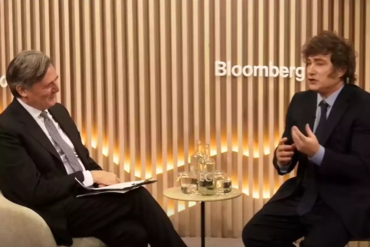 Dos personas en traje conversan en un set de Bloomberg con un fondo de paneles de madera y una mesa con botellas de agua.