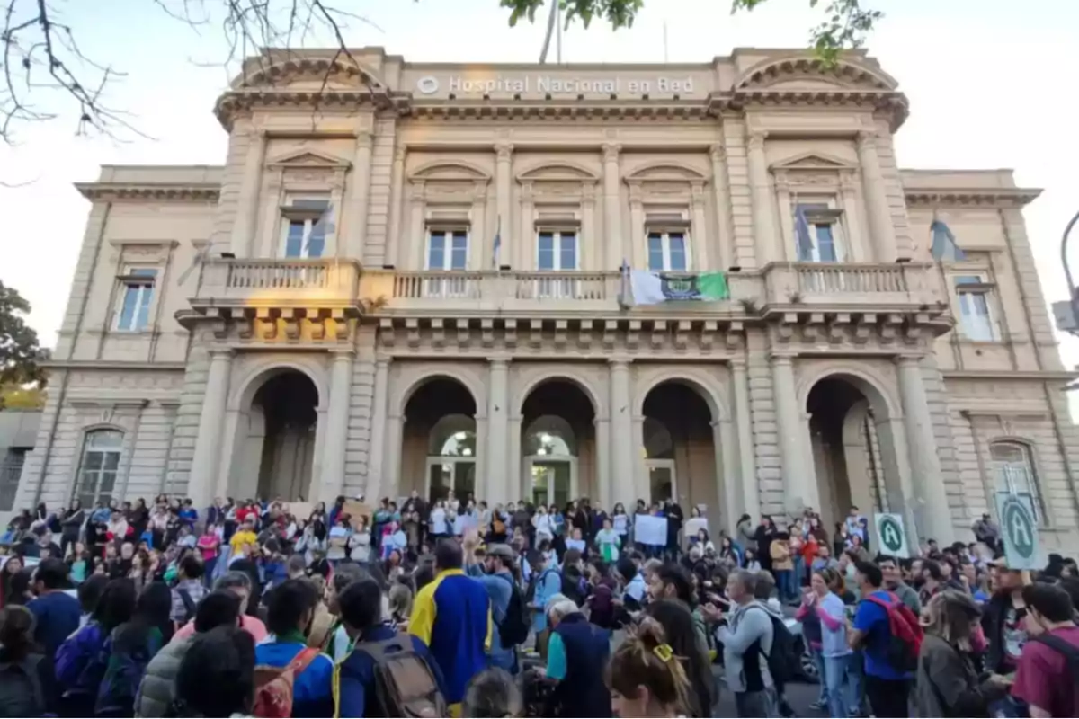 Un grupo de personas se reúne frente a un edificio histórico con columnas y balcones, que tiene un cartel que dice 