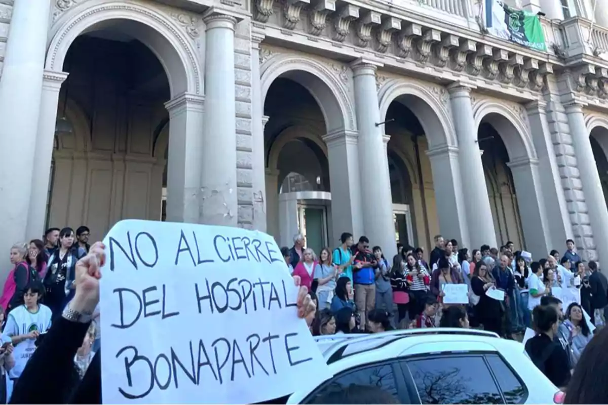 Un grupo de personas se manifiesta frente a un edificio con columnas, sosteniendo carteles que dicen 